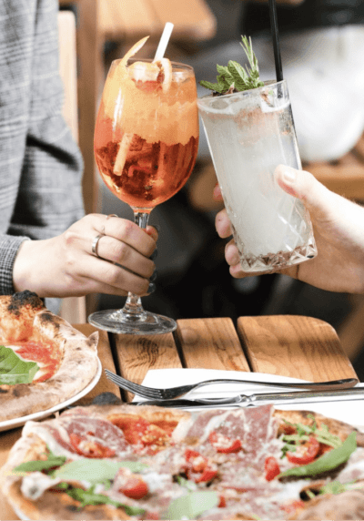 Two people clink cocktail glasses. Two large pizzas are on the table below them.
