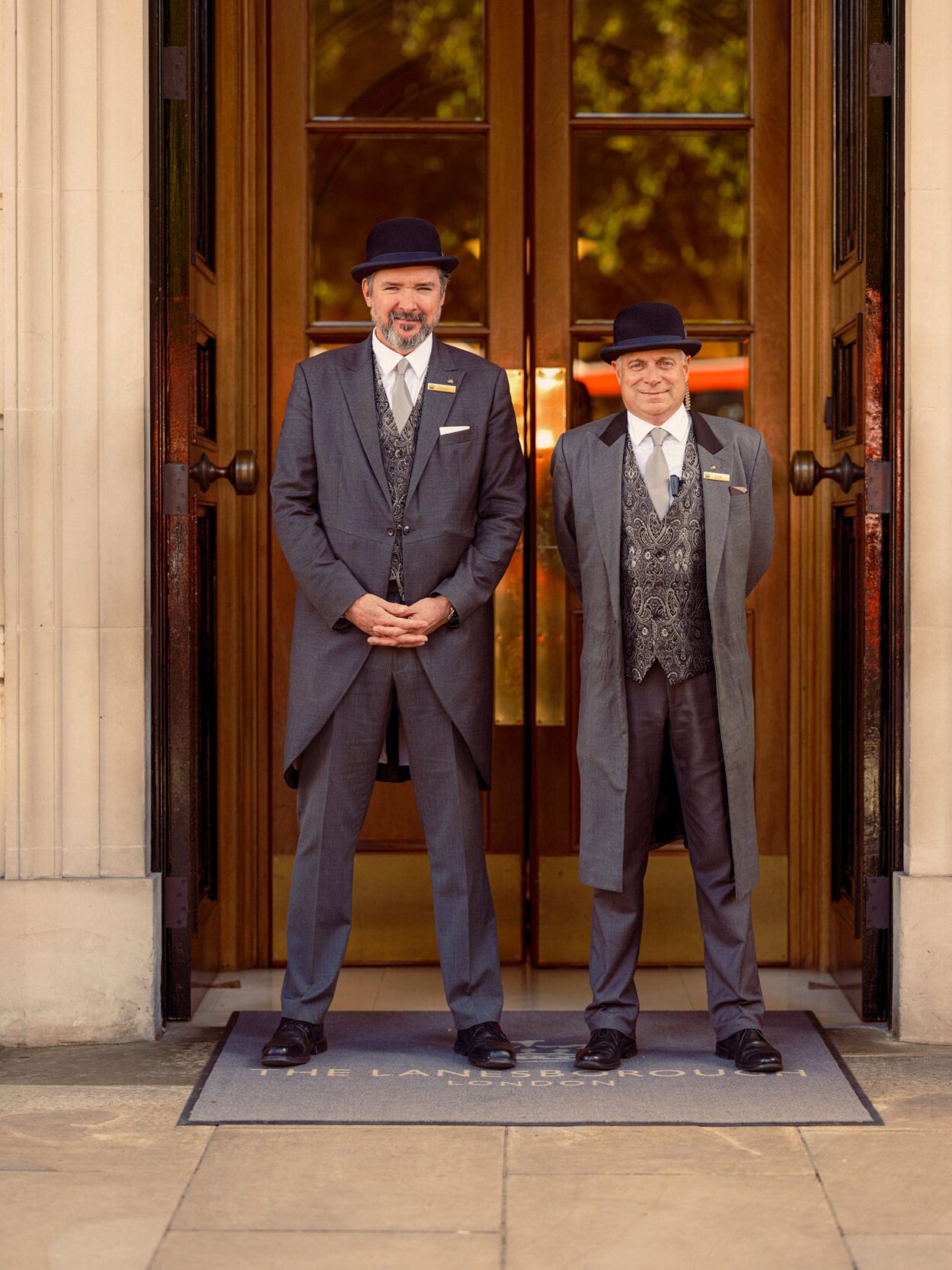 Two doorman, one taller than the other, stand by the entrance to a building. They are wearing top hats and formal attire.
